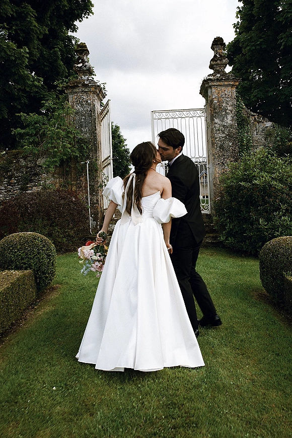 Wedding kiss portrait of bride and groom kissing, bride holding bouquet in strapless gown with puff sleeves by iron gate in garden lawn