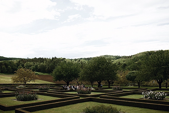 Garden wedding ceremony with wedding guests seated in white chairs along the lawn, framed by manicured hedges and rolling hills under cloudy sky