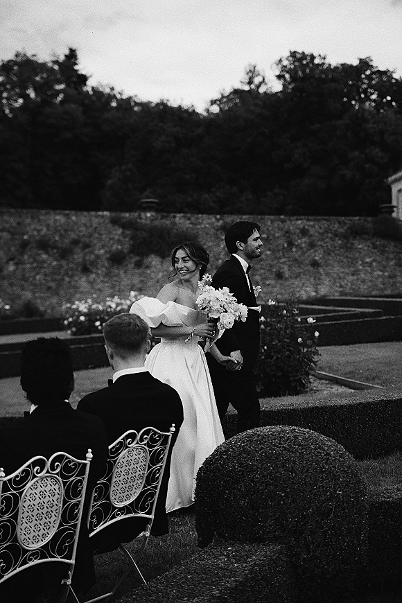 Wedding recessional as bride and groom walk the aisle holding hands, bouquet and tuxedo in a formal garden with chairs and hedges