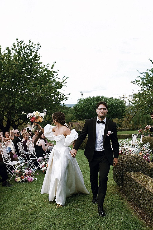 Wedding recessional as bride and groom walking aisle hand in hand, bouquet raised overhead, guests cheering on a garden lawn with white chairs