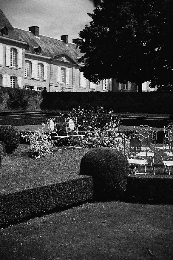 Ceremony setup with white metal chairs lining a floral meadow aisle in a formal garden, with hedges, a large tree, and historic facade beyond