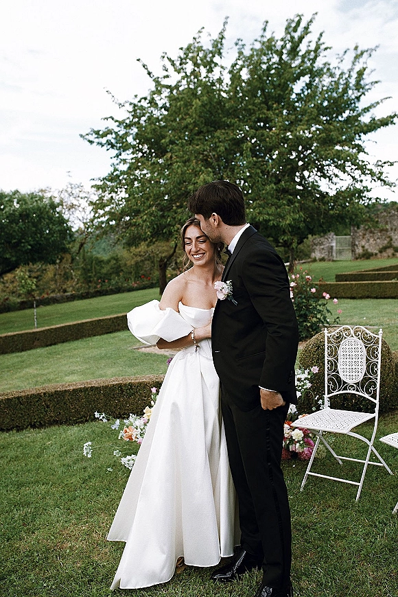 Couple portrait of bride and groom embrace, forehead kiss, in strapless dress and black tuxedo on a garden lawn by hedges
