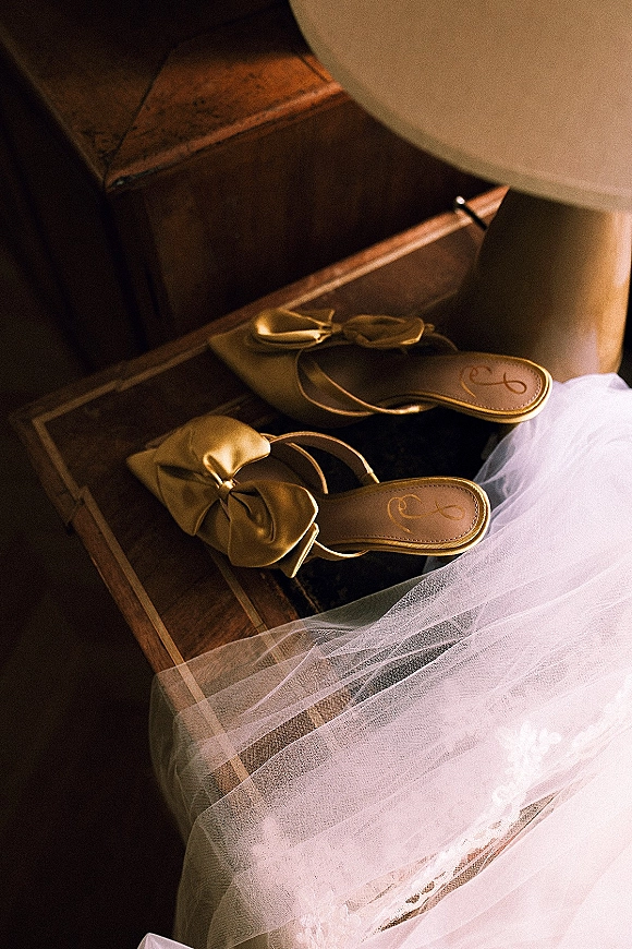 Bridal shoes with bow wedding heels in ivory satin, pointed toes beside a tulle veil on wooden stairs by a lamp base