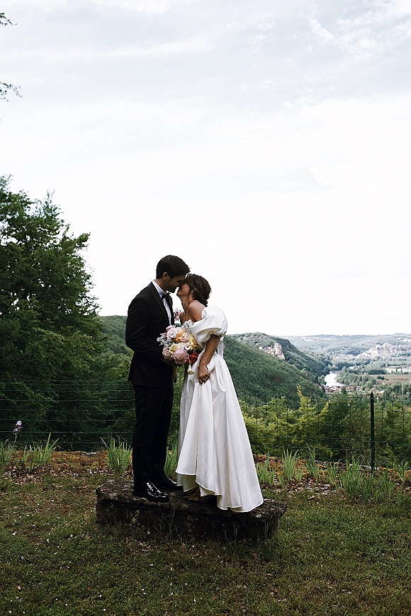 Wedding kiss portrait of bride and groom kissing, her bouquet visible, on a stone platform overlook with river valley and cloudy mountains behind