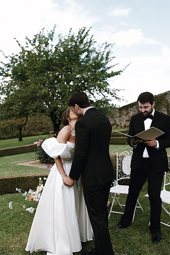 Ceremony kiss at an outdoor wedding ceremony as the bride in an off-shoulder gown and groom in black tuxedo kiss on a garden lawn