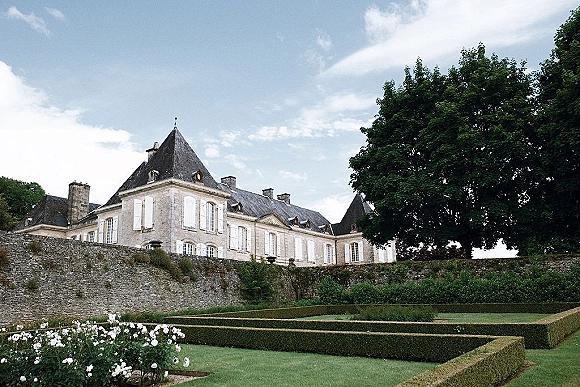 Wedding venue exterior with formal hedges and garden flowers beside a stone wall, facing a historic stone manor under a cloudy sky
