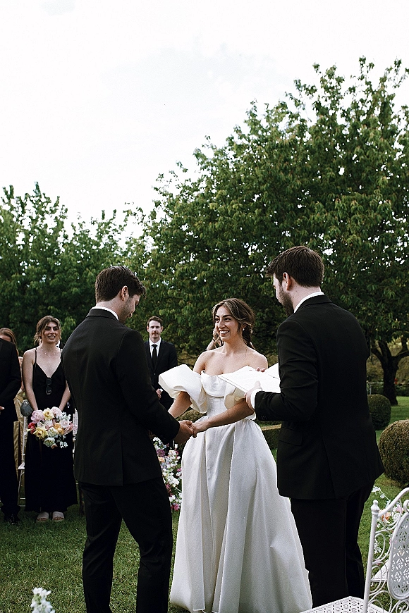 Wedding vows as bride and groom hold hands while officiant reads from a book, pastel bouquet accents in a garden lawn ceremony