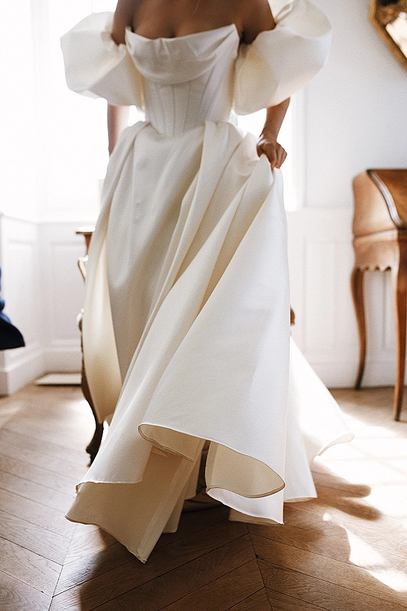 Wedding dress with off the shoulder neckline, puff sleeves, and satin corset bodice, full skirt and train in a sunlit paneled room