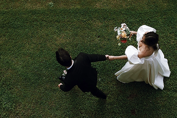 Couple portrait from above of bride in a puff sleeve gown holding a colorful bouquet, walking hand in hand with groom in black tux on grass lawn