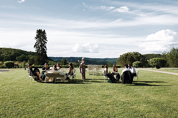 Wedding cocktail hour with guests mingling around outdoor lounge chairs and a sofa, holding cocktail glasses on a lawn with rolling hills