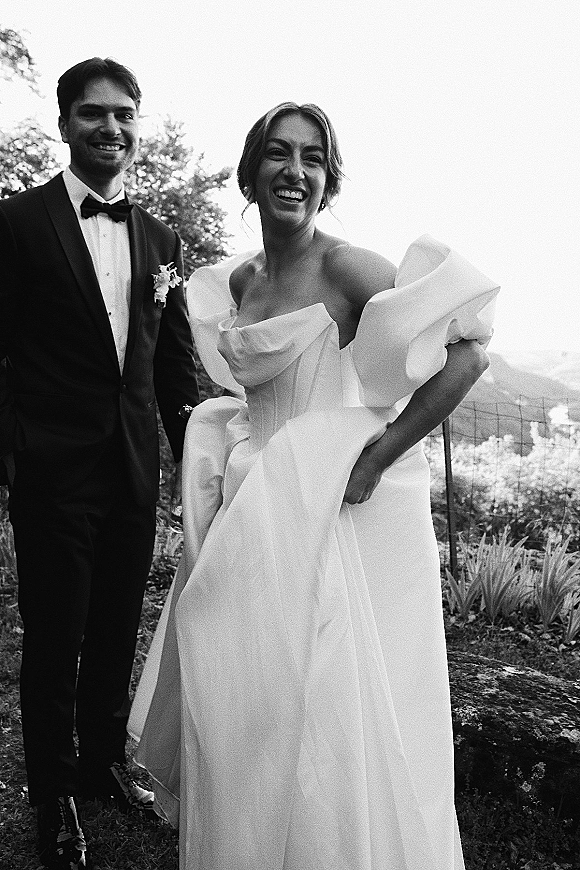 Couple portrait in black and white, bride laughing in off-shoulder wedding dress beside groom in tuxedo with bow tie in a vineyard garden setting