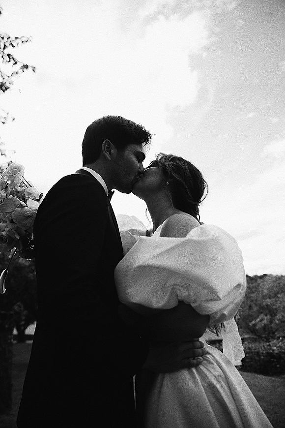 Wedding kiss portrait of bride and groom kissing, her puff sleeve gown and bouquet visible, under dramatic clouds on a hillside