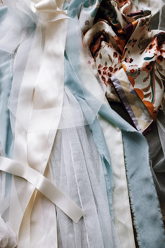Wedding ribbon flatlay with silk ribbon styling, sheer tulle and fabric swatches arranged on a textured fabric surface with a floral print scarf