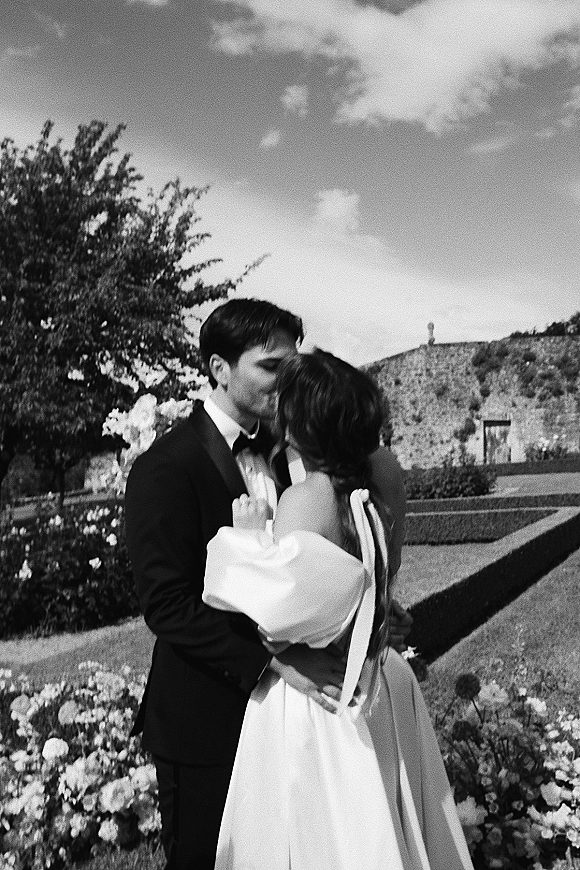 Wedding kiss portrait of bride and groom kissing in a hedge maze garden, her off-the-shoulder dress and ribbon ties beside stone wall