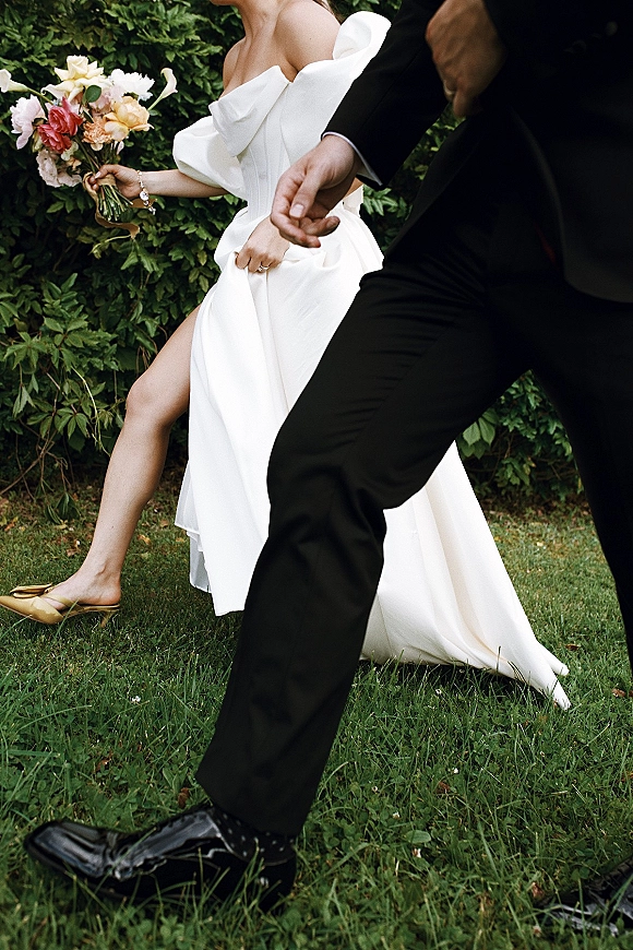 Couple portrait of bride and groom walking on a garden lawn, bride holding a ribbon-wrapped bouquet in an off the shoulder dress with slit