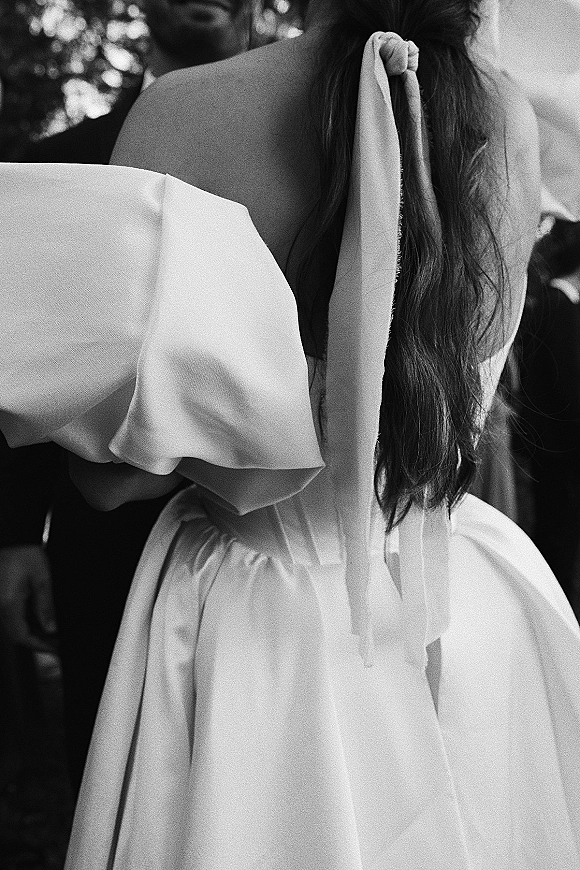 Wedding couple portrait of bride and groom embrace, her off-the-shoulder dress and bow ribbon visible against soft tree bokeh outdoors