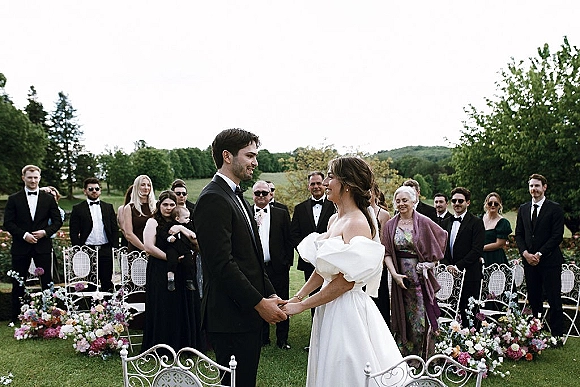 Ceremony moment at an outdoor wedding ceremony with bride and groom holding hands, guests standing by colorful aisle florals on a grassy lawn with trees and hills