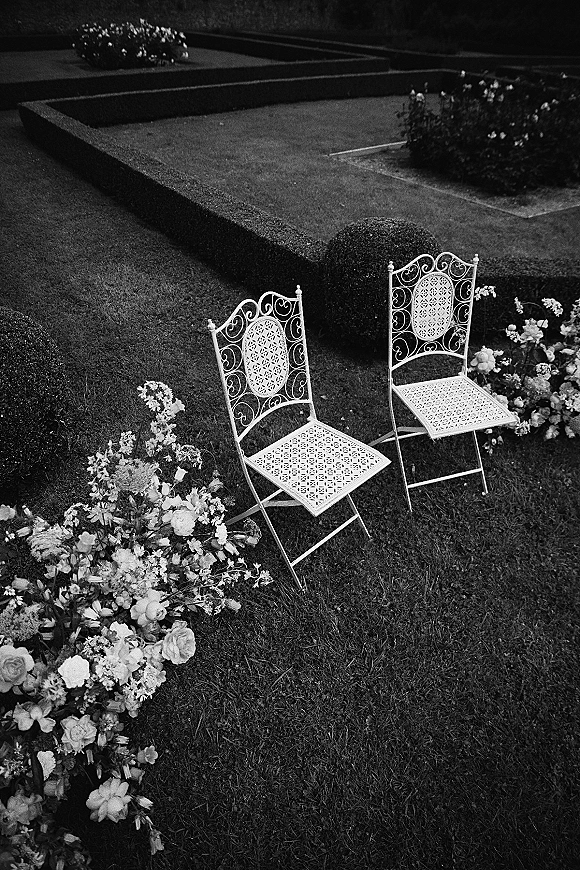 Ceremony seating with white wrought iron chairs and rose greenery ground florals on a lawn bordered by manicured hedges