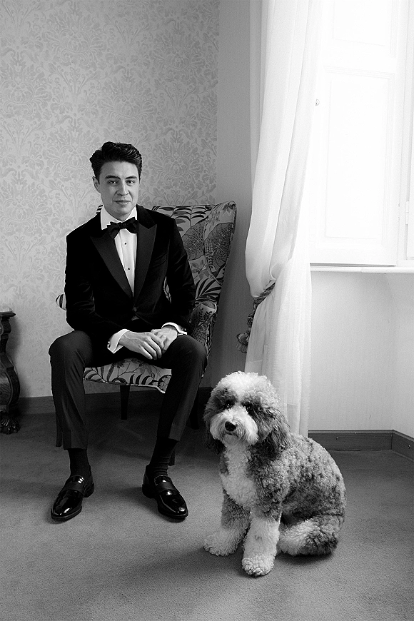 Groom portrait of a groom in tuxedo seated in a patterned armchair with his dog, black bow tie crisp under soft window light