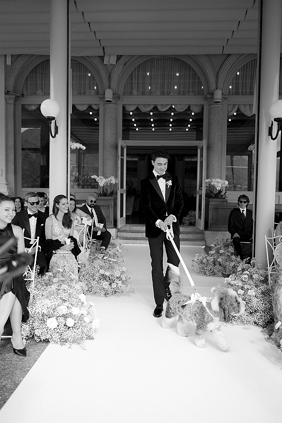 Wedding processional with a dog ring bearer as the groom in a black tuxedo leads him on a leash down a white aisle runner by roses