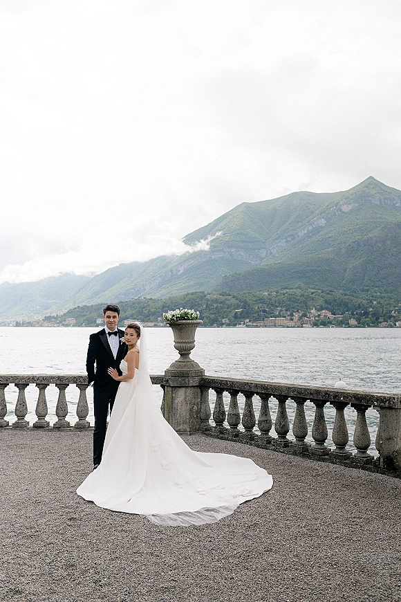 Couple portrait of bride and groom posing on a stone terrace, her strapless gown and veil flowing, with mountain lake backdrop under clouds