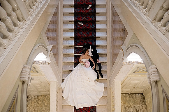 Couple portrait of bride and groom on stairs, bride in strapless gown holding bouquet beside groom in tux on a grand marble staircase