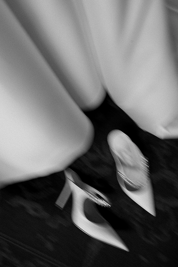 Bridal shoes in a wedding shoes flatlay, pointed-toe heels with rhinestone embellishment beside soft bridal gown fabric on a dark floor