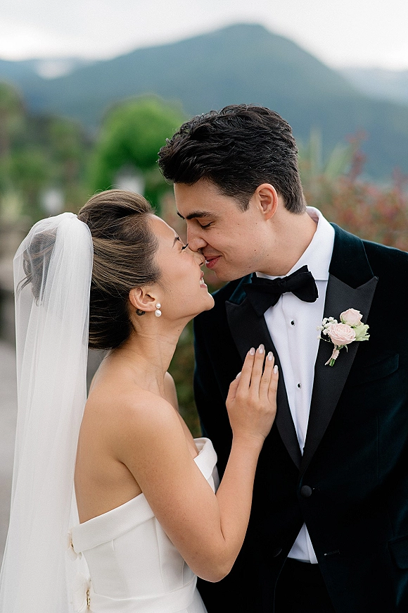 Wedding kiss portrait of bride and groom kissing, her veil and bouquet against his black tuxedo, with mountains and sky behind