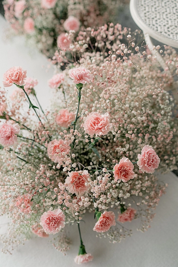 Wedding florals with a pink carnation centerpiece and baby's breath nestled in greenery on a white tablecloth against a soft neutral backdrop