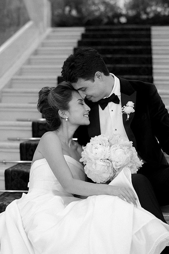 Wedding couple portrait in black and white, bride in strapless dress leaning on tuxedoed groom as he kisses her forehead on staircase steps