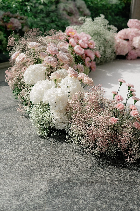 Wedding aisle florals in a ground floral aisle arrangement of roses, hydrangeas, carnations and baby’s breath along a white runner on stone floor