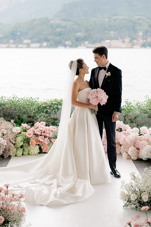 Couple portrait of bride in a strapless satin wedding dress holding a pink rose bouquet beside groom in tux by a lakeside hedge