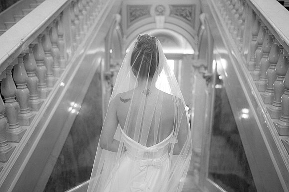 Bridal portrait of a bride from behind in a strapless, backless wedding dress with a wedding veil, descending a grand staircase indoors