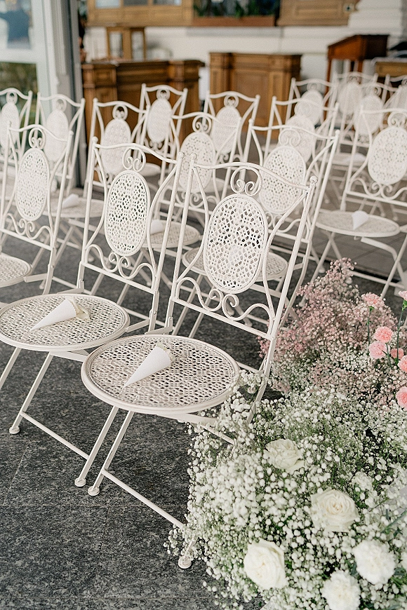 Ceremony seating with white wrought iron wedding chairs lined beside aisle florals of white roses and baby's breath, with confetti cones indoors