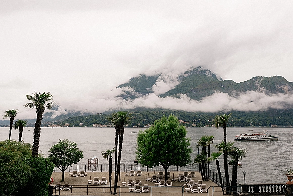 Lakeside landscape with a lake and mountain view, palm trees framing lounge chairs beside a metal fence under low clouds