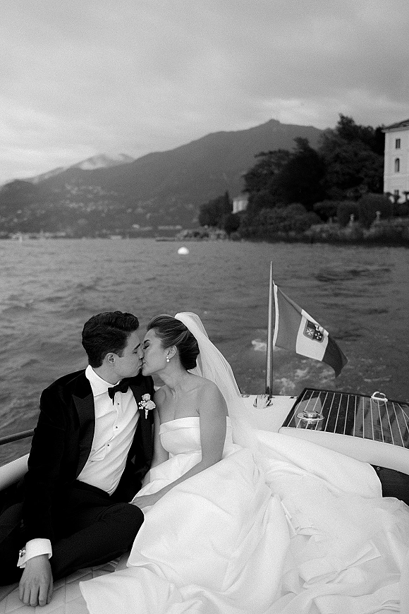Wedding kiss portrait of newlyweds on boat, bride in veil and strapless gown kissing tuxedo groom on a lake with mountains under clouds