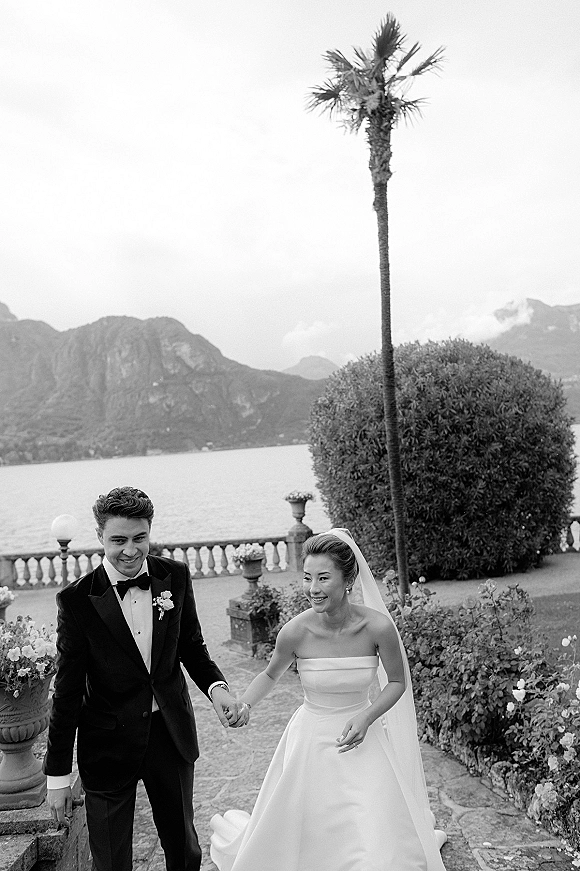 Wedding couple portrait of bride and groom holding hands, laughing on a stone terrace with lake, mountains, and palm tree behind them