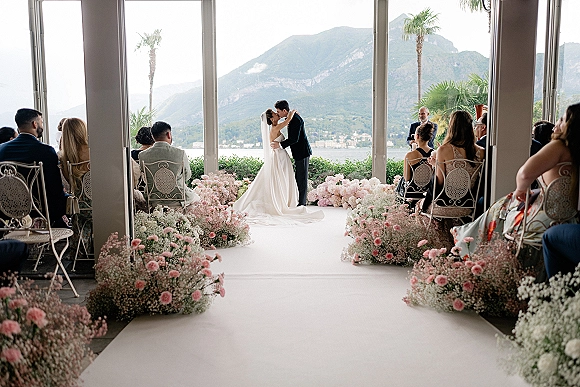 Ceremony kiss as bride in veil and groom in suit embrace on white aisle runner with pink florals, guests and mountain lake view windows behind
