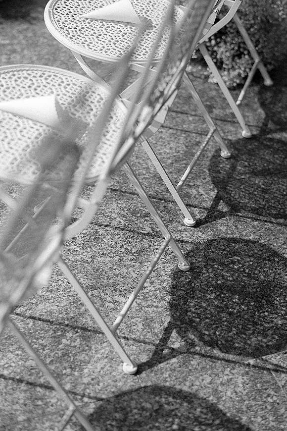 Ceremony seating with wedding ceremony chairs, white metal chairs topped with doilies and paper confetti cones on a stone patio in dappled shade