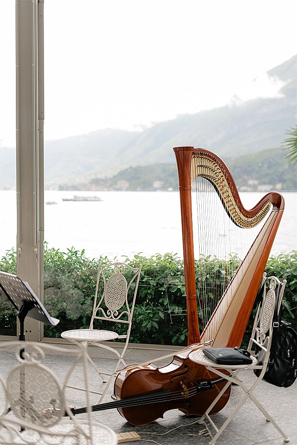 Wedding ceremony music setup with a harp and cello beside a music stand, with instrument cases on a lake-view terrace with mountains