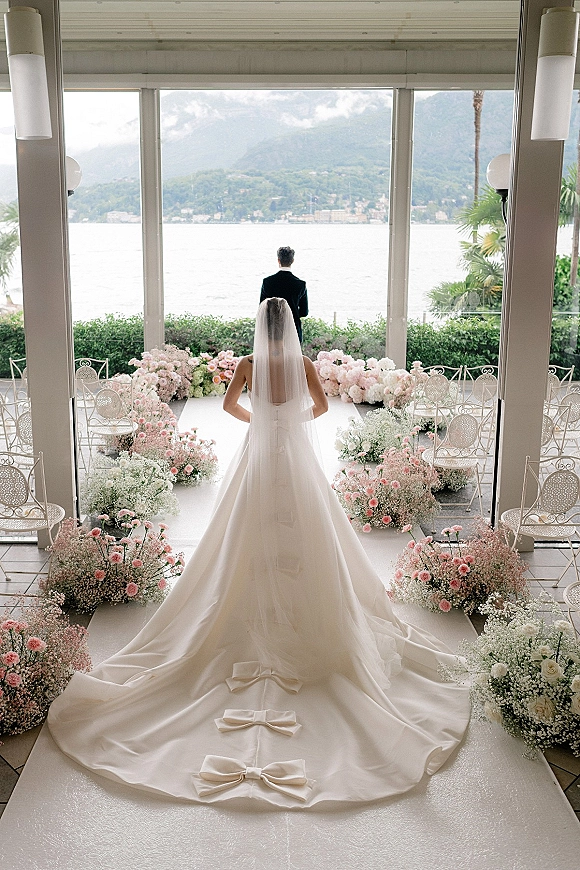 Wedding first look as bride walking to groom, cathedral veil and long train flowing down flower-lined aisle before lake and mountains