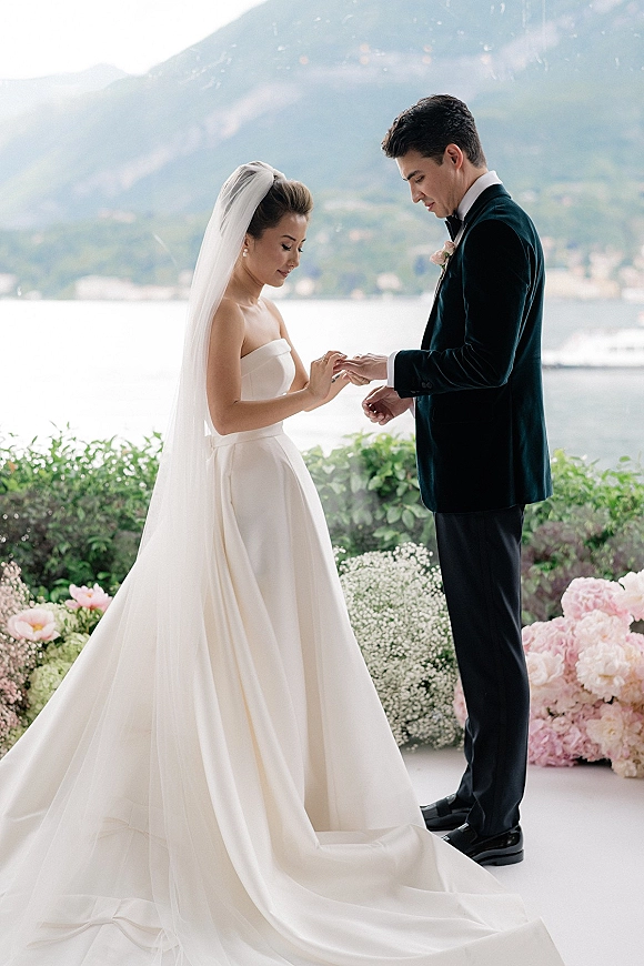 Wedding vows during a ring exchange as the bride in a strapless dress and cathedral veil faces her tuxedoed groom by a mountain lake