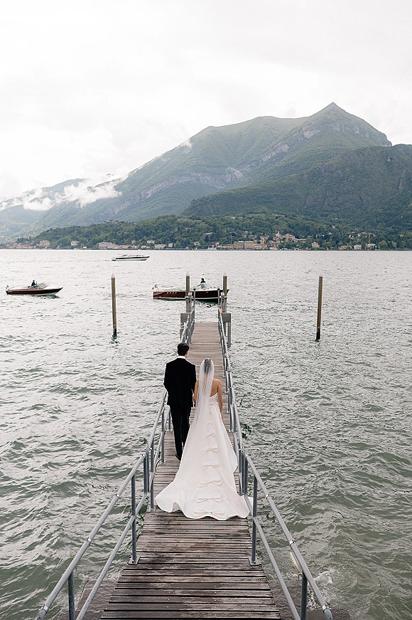 Wedding couple portrait from behind, bride and groom holding hands on a dock with her long train and veil, boats on a mountain lake under clouds