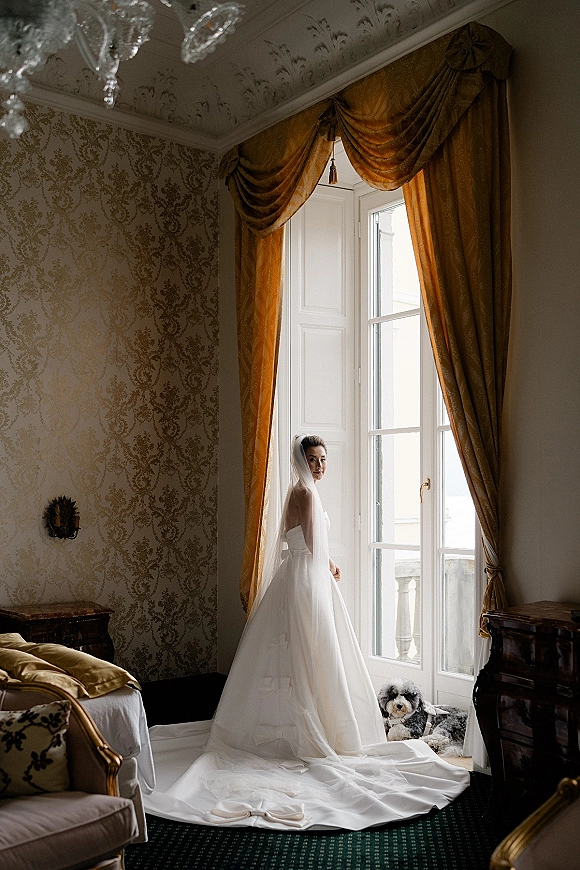 Bridal portrait of bride by window in a strapless ball gown, long veil and train, looking over shoulder beside a small dog in an antique room