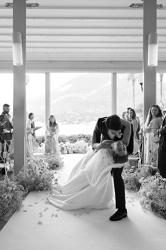 Wedding kiss portrait of groom dipping bride in a long veil on a covered terrace aisle, guests behind, windows framing lake and mountains