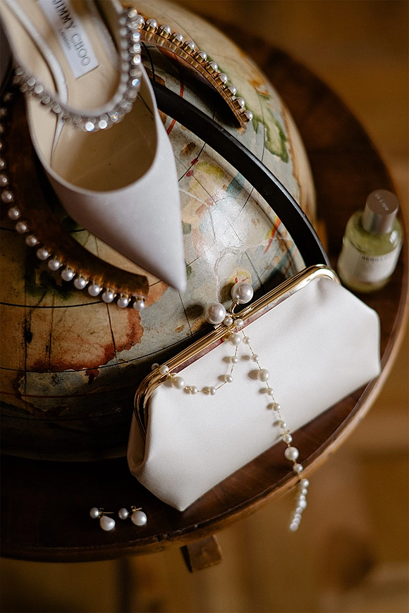 Bridal accessories flatlay with white pointed-toe heels and pearl jewelry, headband, clutch, perfume bottle, and vintage globe on wood surface