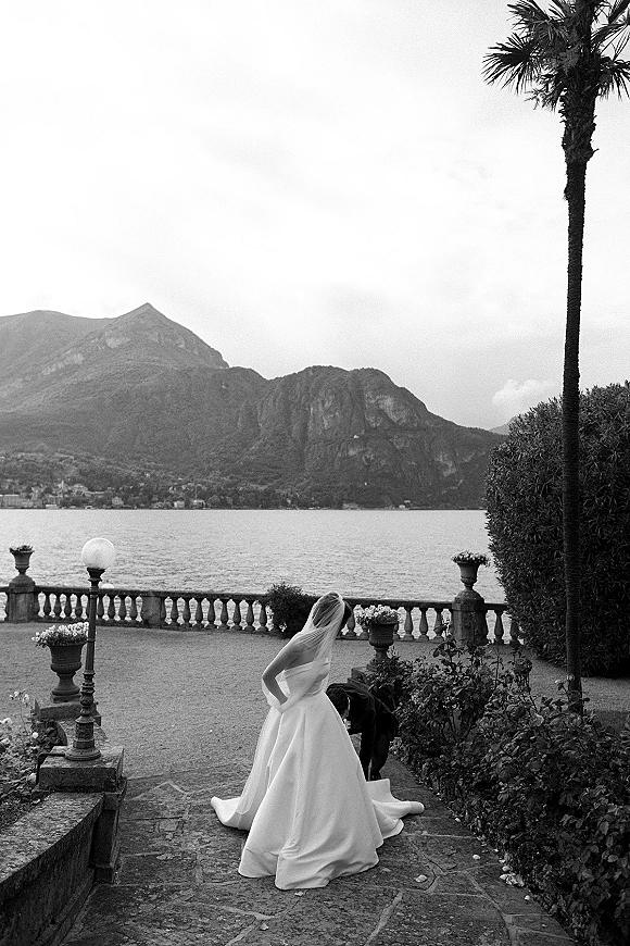Bridal portrait of a bride with veil in a strapless ball gown as the groom adjusts her train on a stone terrace by a lake and mountains