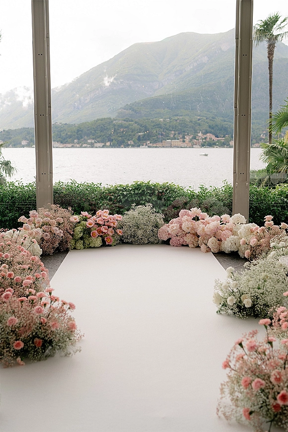 Ceremony aisle design with a white aisle runner bordered by pink roses, hydrangeas, and baby’s breath beside glass walls overlooking a lake and mountains