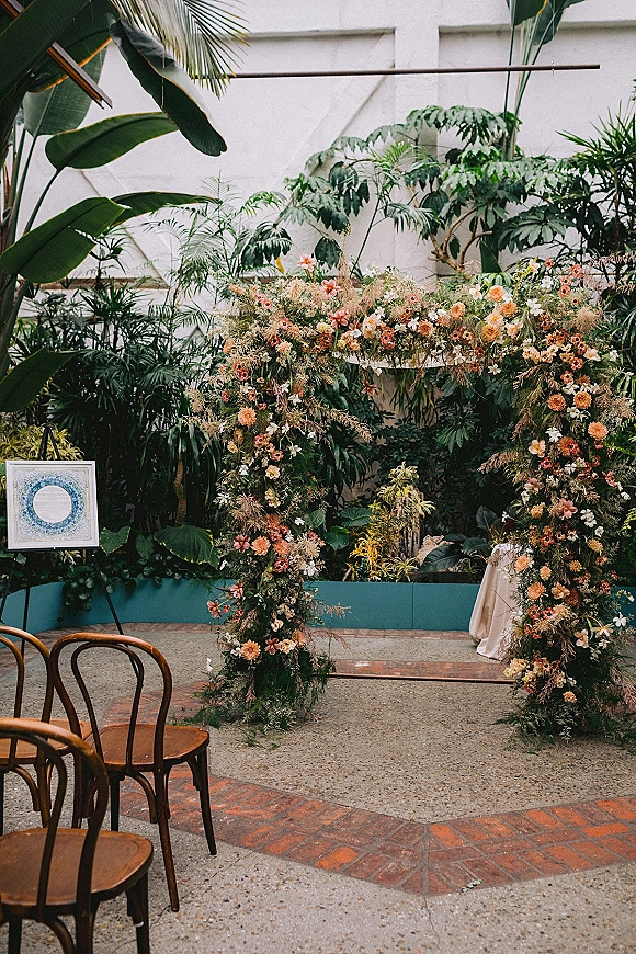 Wedding ceremony backdrop with floral ceremony arch and lush greenery, set in an indoor greenhouse with tropical plants and chairs
