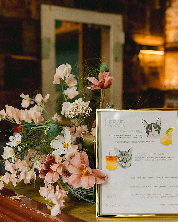 Signature cocktail sign in a gold frame with illustrated cat and drinks, paired with pink-and-white florals on a rustic wood tabletop